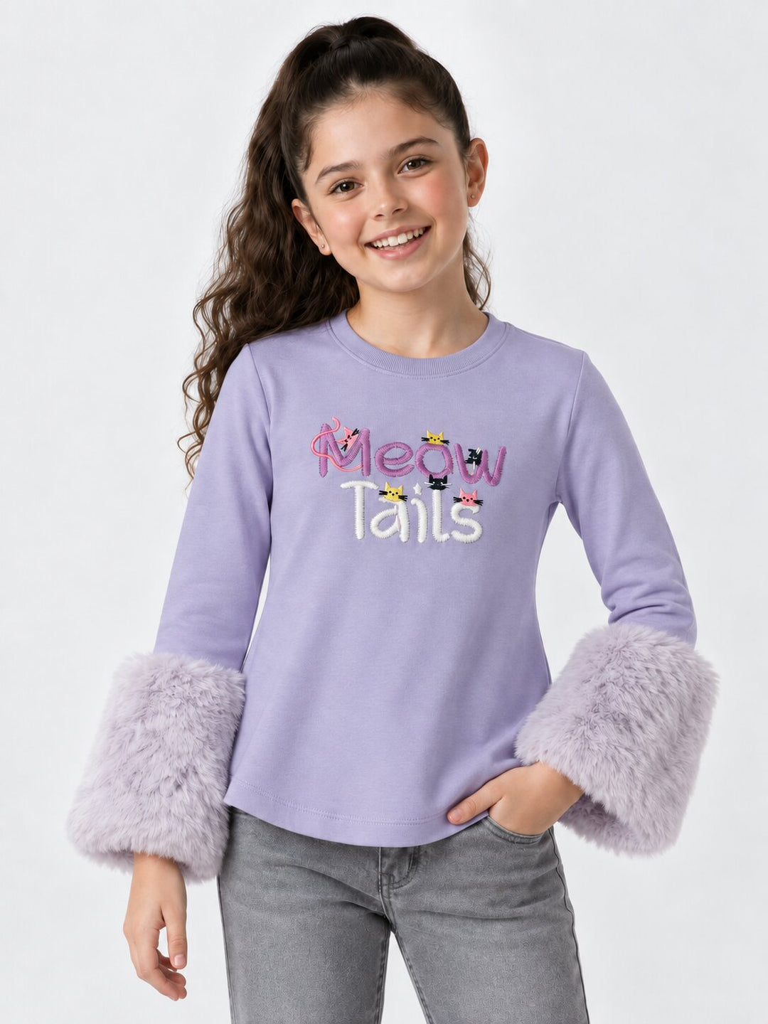 A young girl with long brown hair in a ponytail models a lavender long-sleeve shirt with fuzzy lavender cuffs. The shirt features the text 'Meow Tails' in pink and white with small cartoon bunnies.