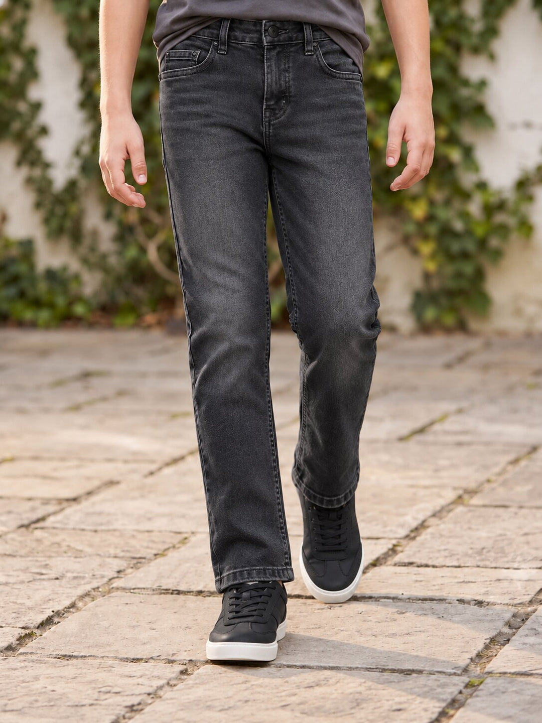 A person wearing dark gray washed denim jeans and black sneakers walking on a stone patio with green ivy in the background.