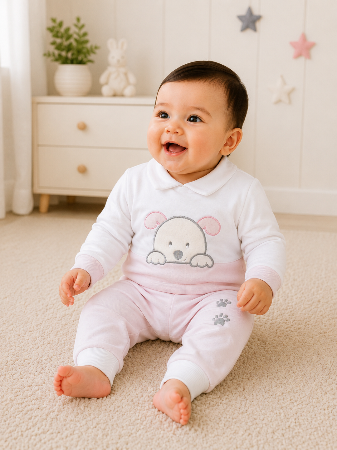 A smiling baby sitting on a beige carpet in a bright nursery, wearing a white long-sleeve top and light pink pants with a cartoon bear face on the chest and small paw prints near the ankles.