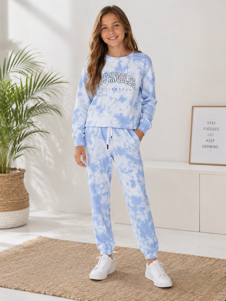 A young woman modeling a light blue and white tie-dye sweatshirt and jogger set with white sneakers in a bright, modern room.
