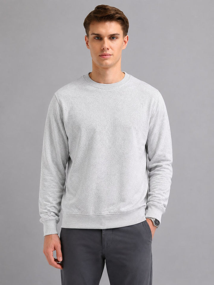 A man wearing a light gray crewneck sweatshirt and dark gray trousers, standing against a solid gray studio background.