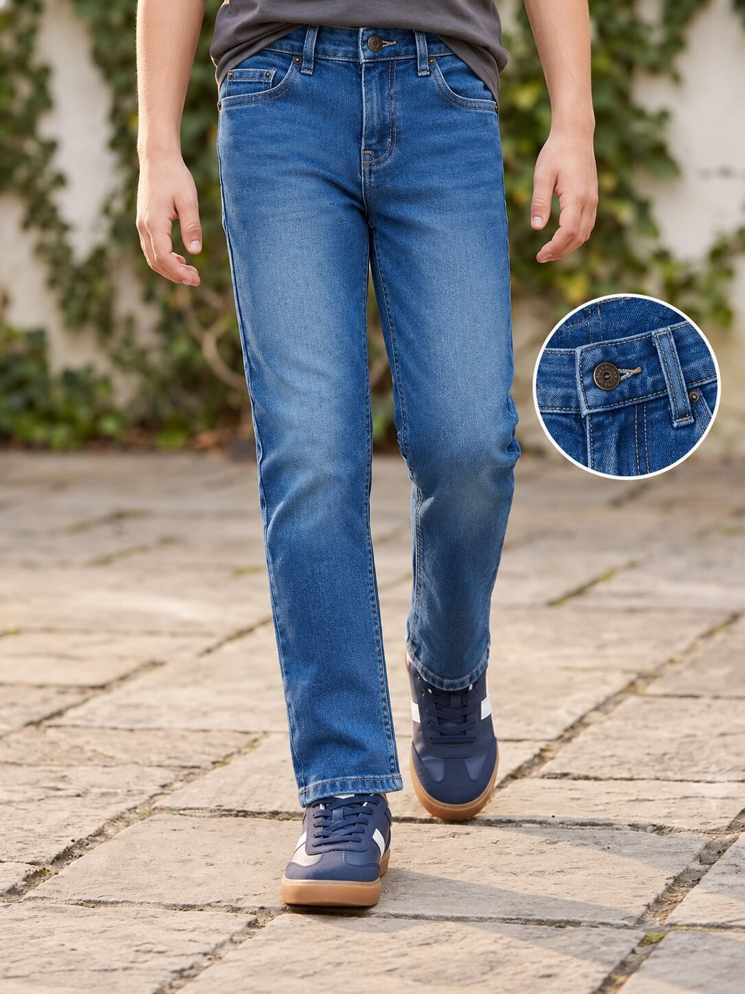 A person wearing medium-wash blue jeans and navy sneakers walking on a stone path, with a circular inset showing a close-up of the jeans' pocket and button detail.