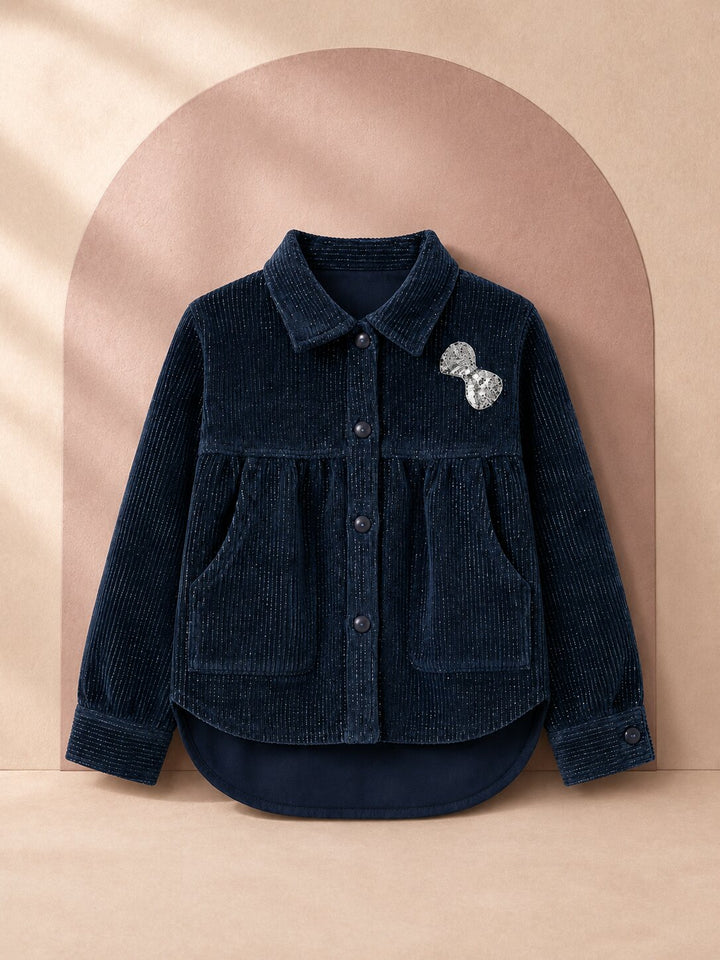 A dark navy blue corduroy button-up shirt jacket with a silver glittery bow pin on the chest, displayed on a beige background with an arched backdrop.