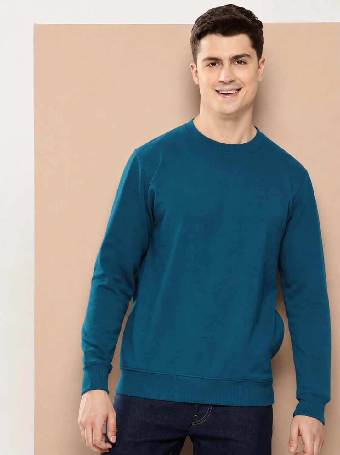 A man wearing a teal blue crewneck sweatshirt and dark jeans, standing against a beige studio backdrop.