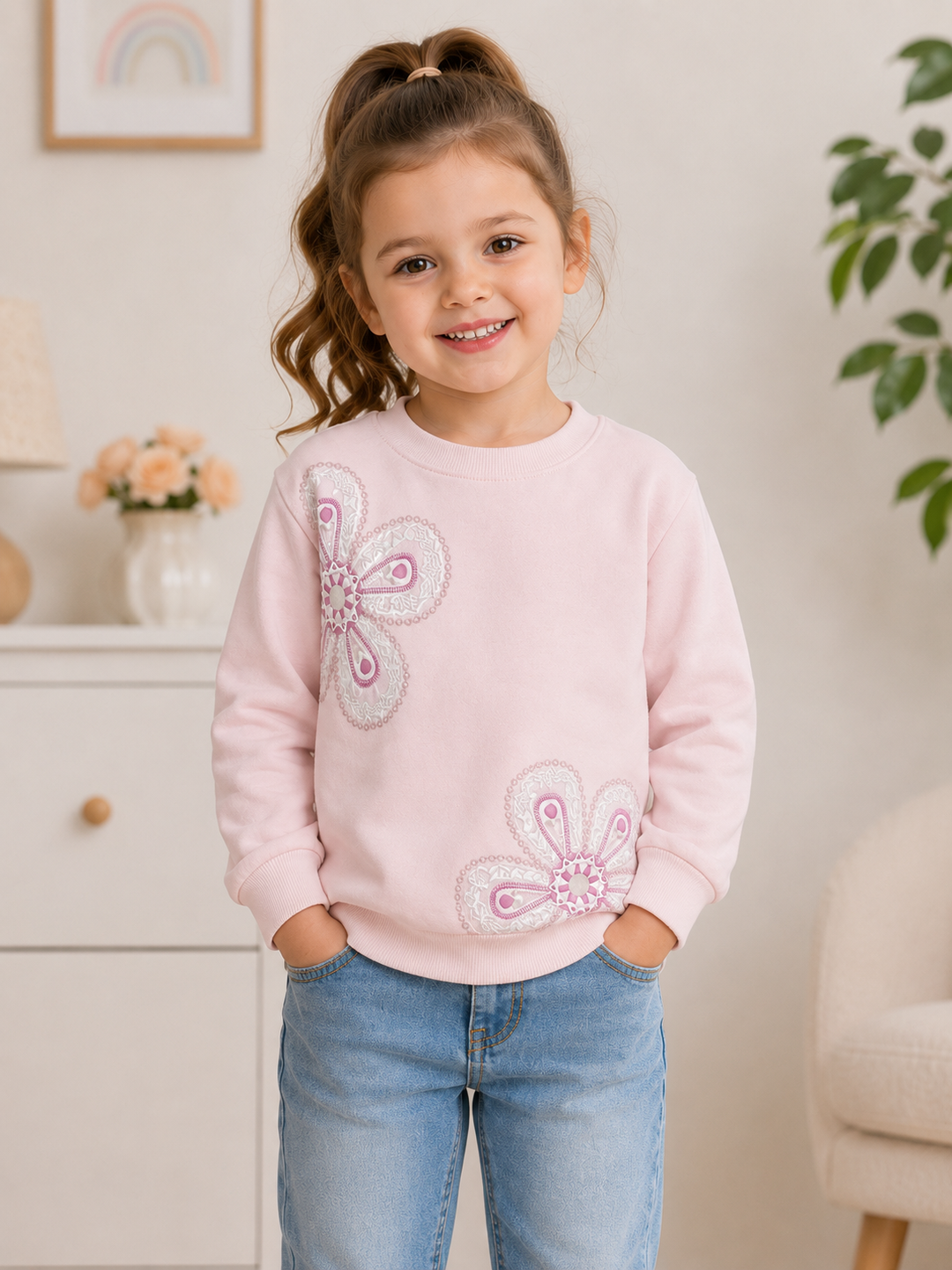 A young girl with brown hair in a high ponytail smiles while wearing a light pink crewneck sweatshirt with two large pink and white butterfly appliques, paired with light blue jeans, standing in a brightly lit room.