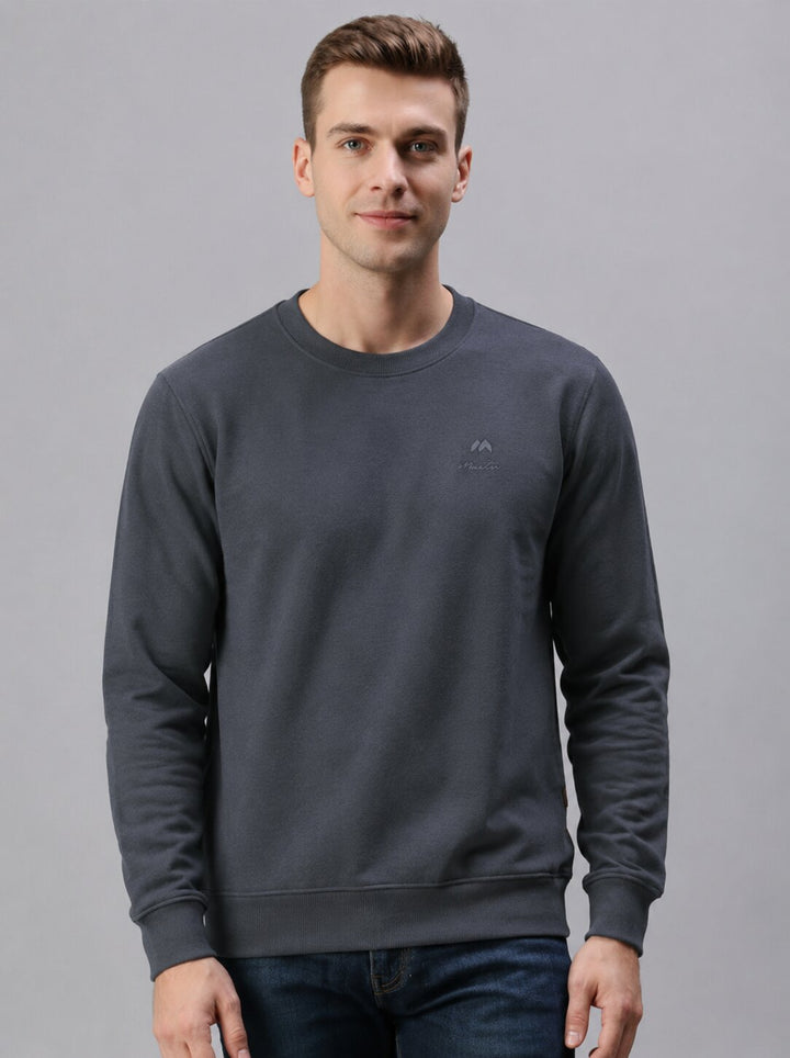 A man wearing a dark gray crewneck sweatshirt with a small tonal logo on the left chest, paired with dark blue jeans, against a solid gray studio background.