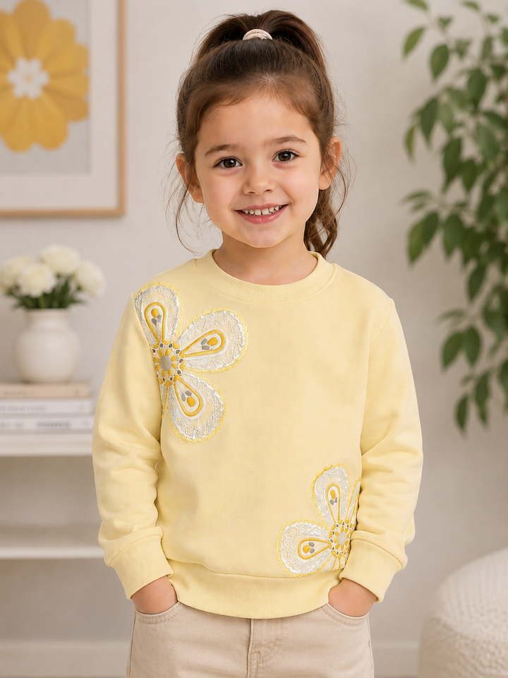 A young girl with her hair in a high ponytail smiles while wearing a pale yellow crewneck sweatshirt with two large white and yellow butterfly appliques on the front, paired with beige pants, in a brightly lit indoor setting.