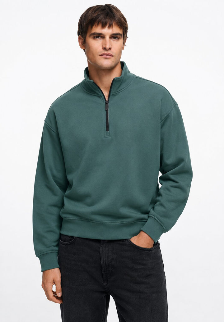 A man wearing a dark green quarter-zip sweatshirt and black jeans, standing against a plain white studio background.