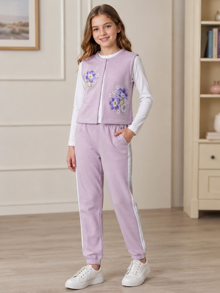 A young girl stands in a room wearing a lavender two-piece tracksuit with a floral embroidered vest over a white long-sleeve shirt and matching lavender jogger pants with white side stripes. She is wearing white sneakers.
