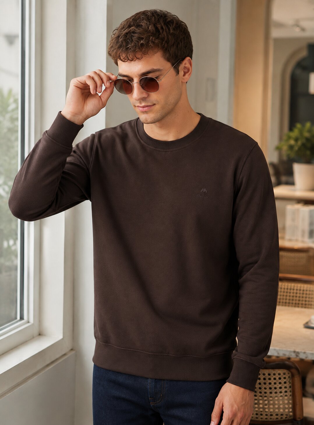 A man wearing a dark brown crewneck sweatshirt and blue jeans, standing in a bright indoor setting near a window and adjusting his sunglasses.