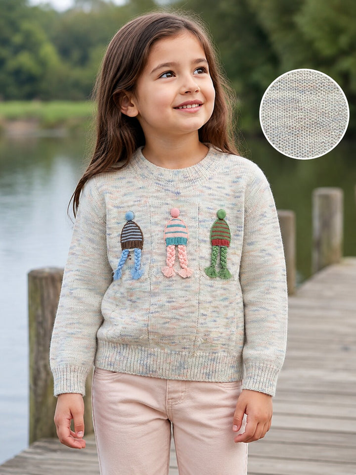 A young girl stands on a wooden dock by the water wearing a light heathered knit sweater with three colorful knit figures on the front. A circular inset in the upper right shows a close-up of the knit texture.