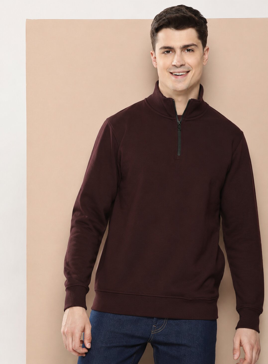 A man wearing a dark brown quarter-zip sweatshirt and blue jeans, standing against a beige background.