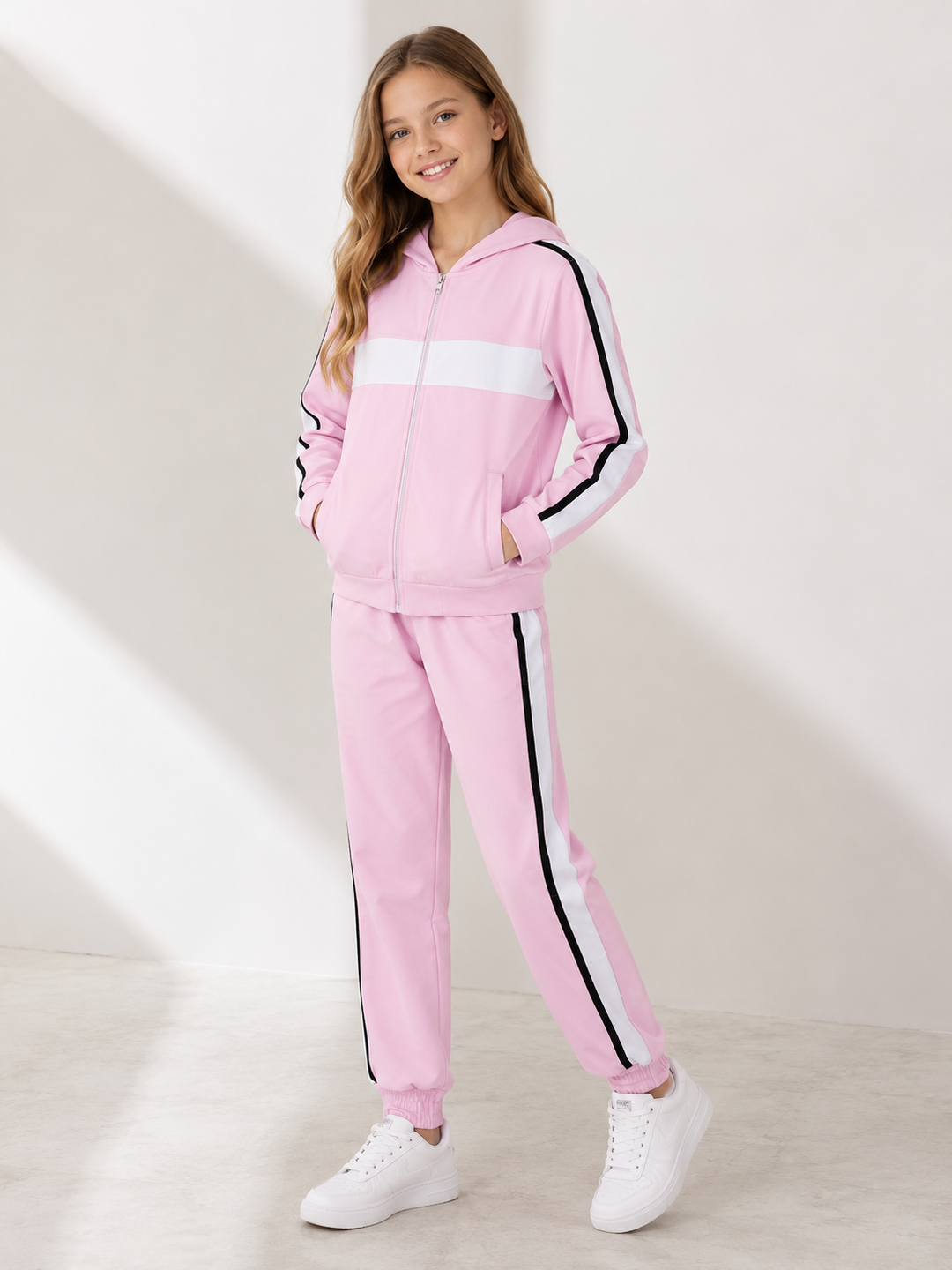 A young girl modeling a pink zip-up tracksuit with white and black stripe details, paired with white sneakers, in a brightly lit studio setting.