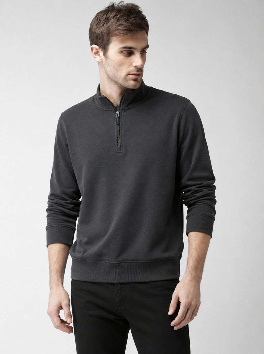A man wearing a dark charcoal gray quarter-zip pullover sweatshirt and black pants, standing against a light gray studio background.