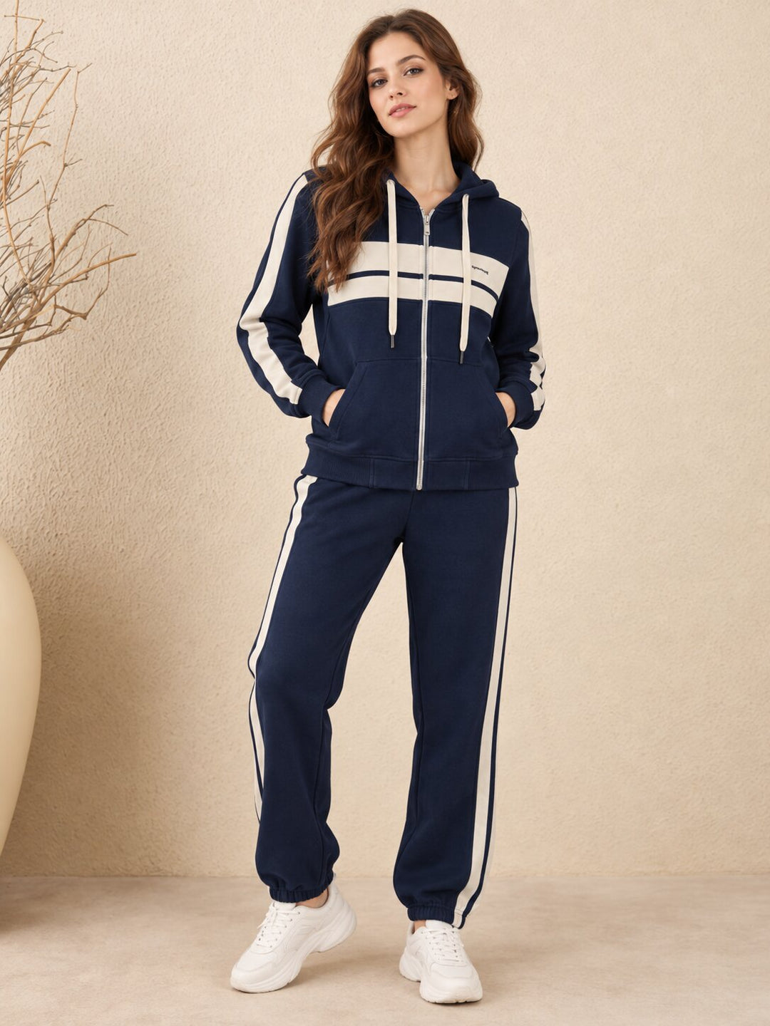 A woman modeling a navy blue tracksuit with white stripes, featuring a zip-up hoodie and matching jogger pants, paired with white sneakers against a textured beige background.