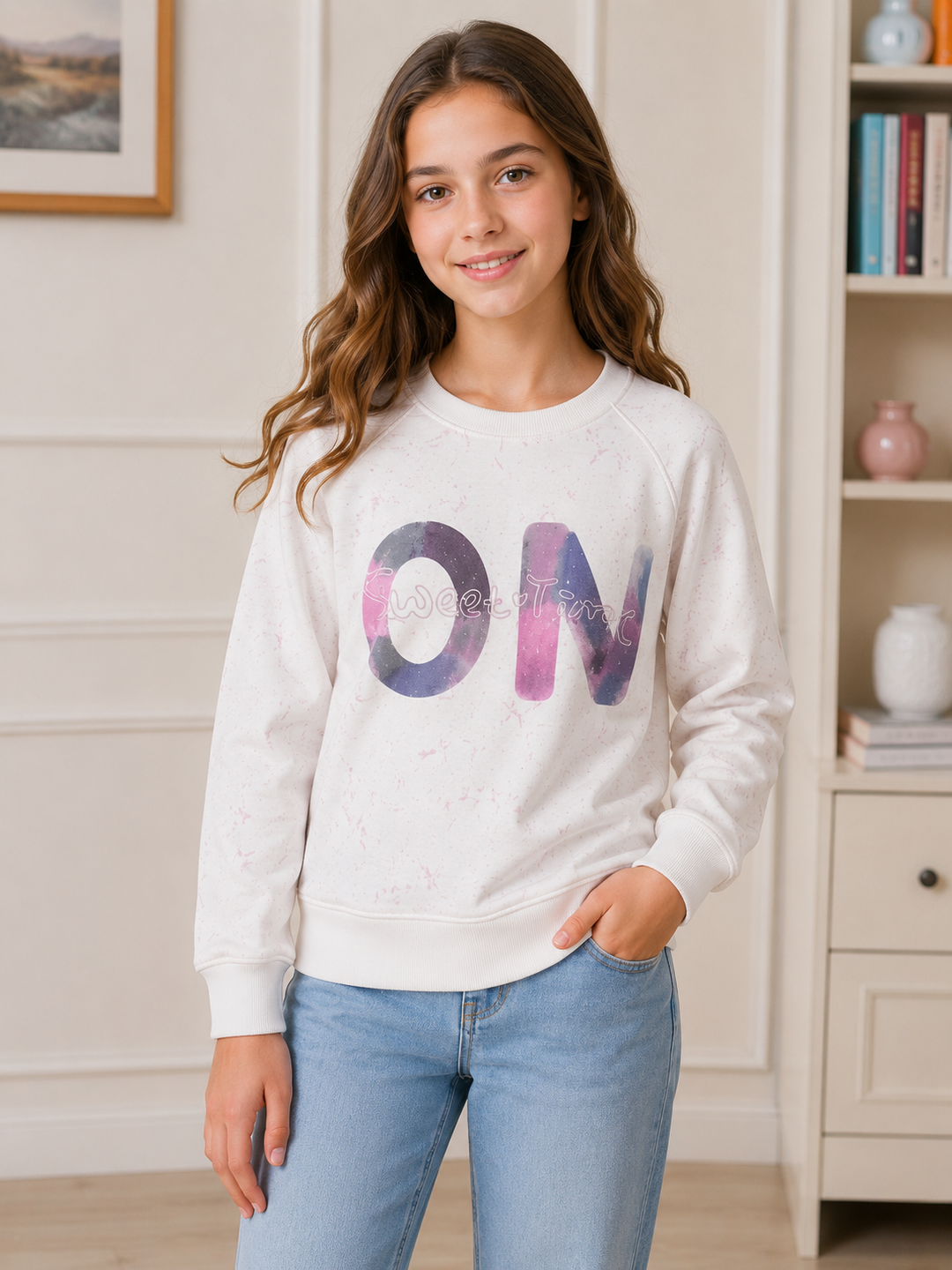 A young girl with long brown hair stands indoors wearing a white crewneck sweatshirt with a large watercolor-style 'ON' graphic in purple and pink on the front, paired with light blue jeans.