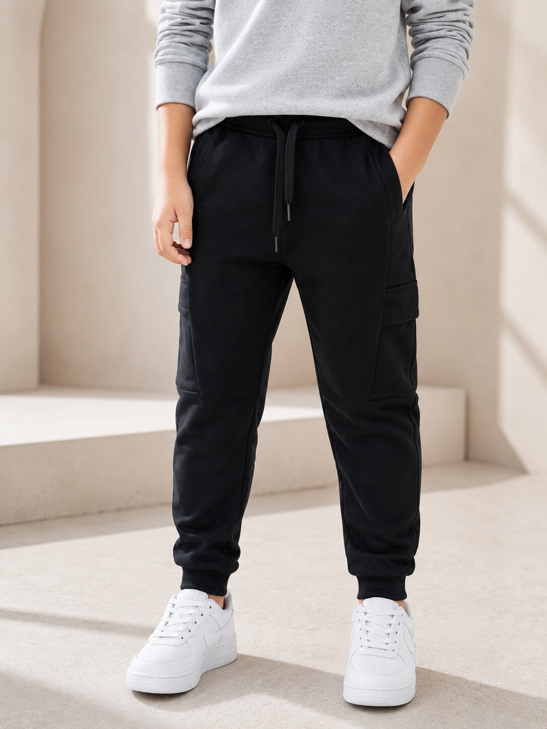 A person wearing black cargo jogger pants with a drawstring waist, paired with a light gray sweatshirt and white sneakers, standing in a minimalist beige architectural setting.