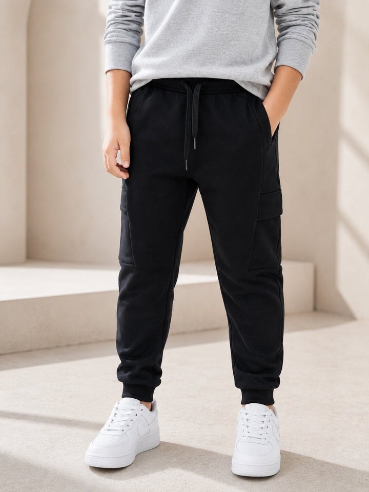 A person wearing black cargo jogger pants with a drawstring waist, paired with a light gray sweatshirt and white sneakers, standing in a minimalist beige architectural setting.