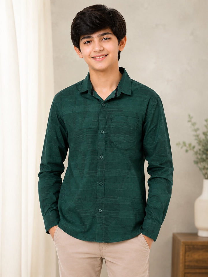 A young man wearing a dark green long-sleeve button-up shirt with a subtle grid pattern, paired with beige trousers, standing in a softly lit indoor setting.