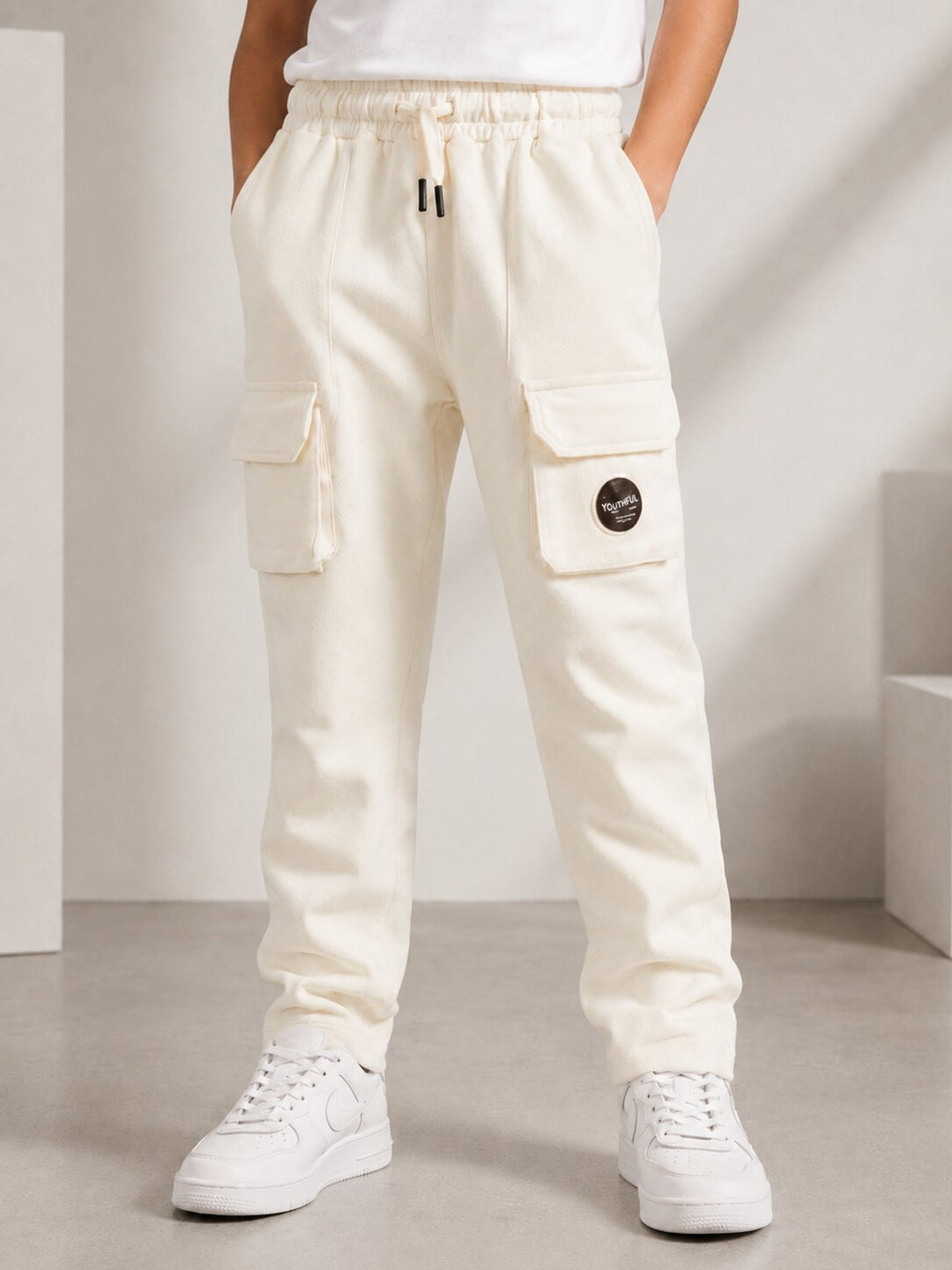 A person wearing off-white cargo jogger pants with side flap pockets, a drawstring waist, and white sneakers, standing in a minimalist studio setting.