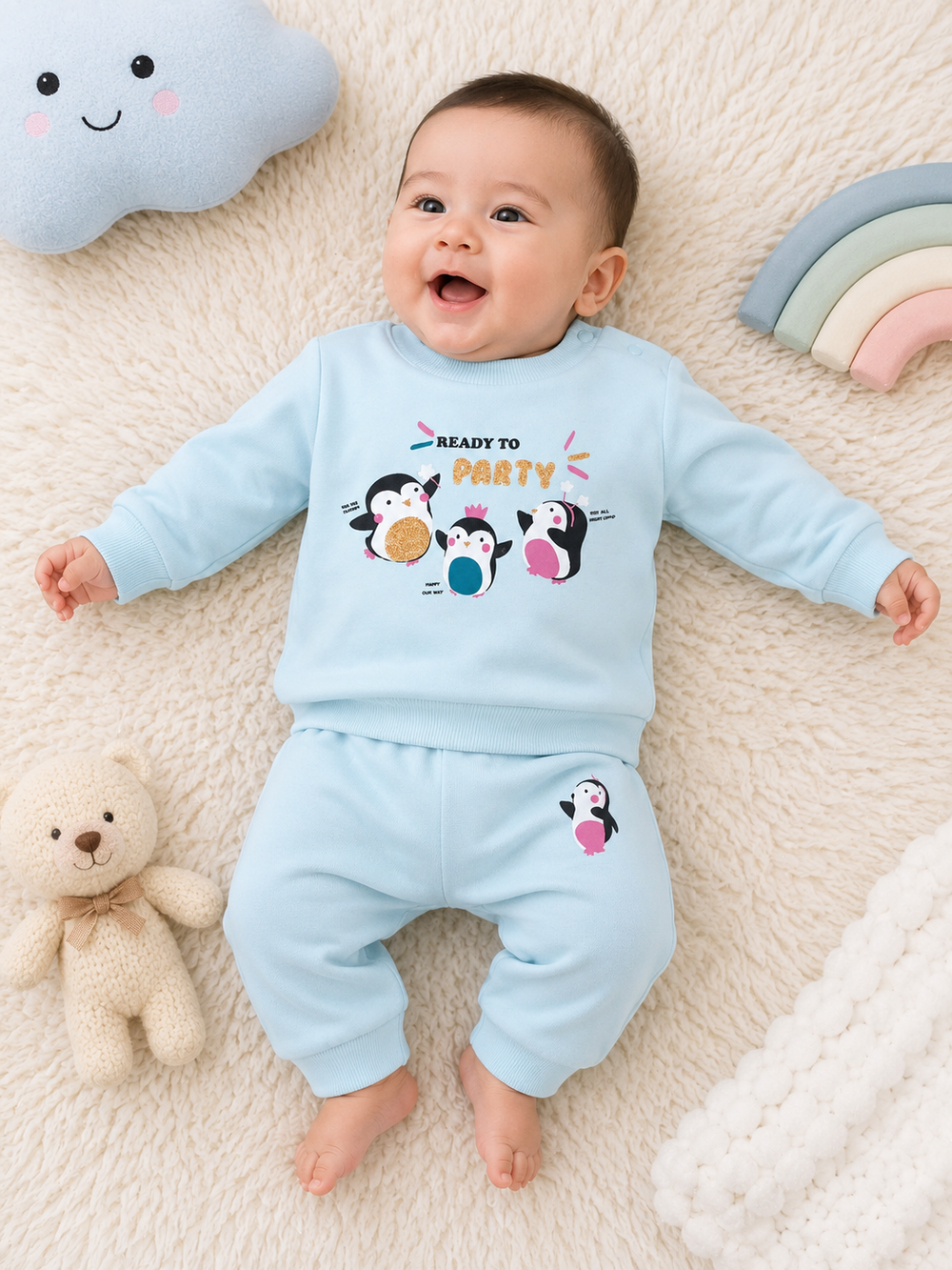 A baby lying on a cream faux-fur blanket wearing a light blue two-piece outfit with a penguin party graphic on the top and a small penguin print on the pants. The scene includes a plush cloud toy, a teddy bear, and a pastel rainbow in the background.