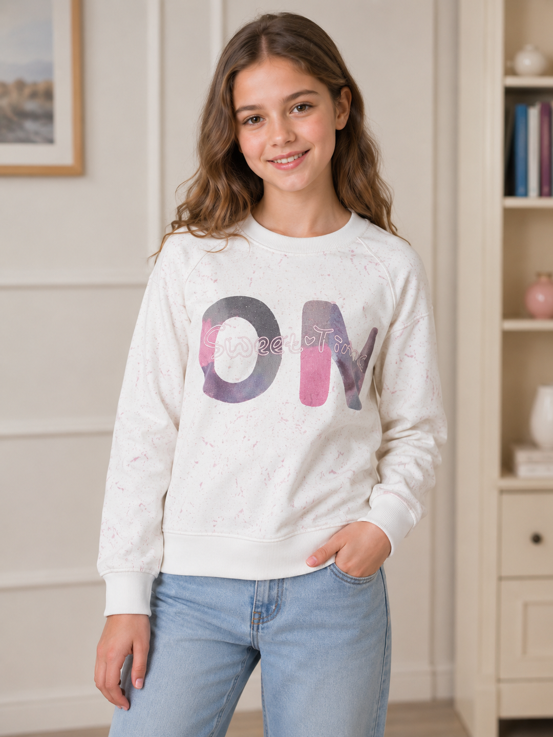 A young girl with wavy brown hair smiling, wearing a white crewneck sweatshirt with a colorful 'ON' graphic and light blue jeans, standing in a brightly lit room with a bookshelf in the background.