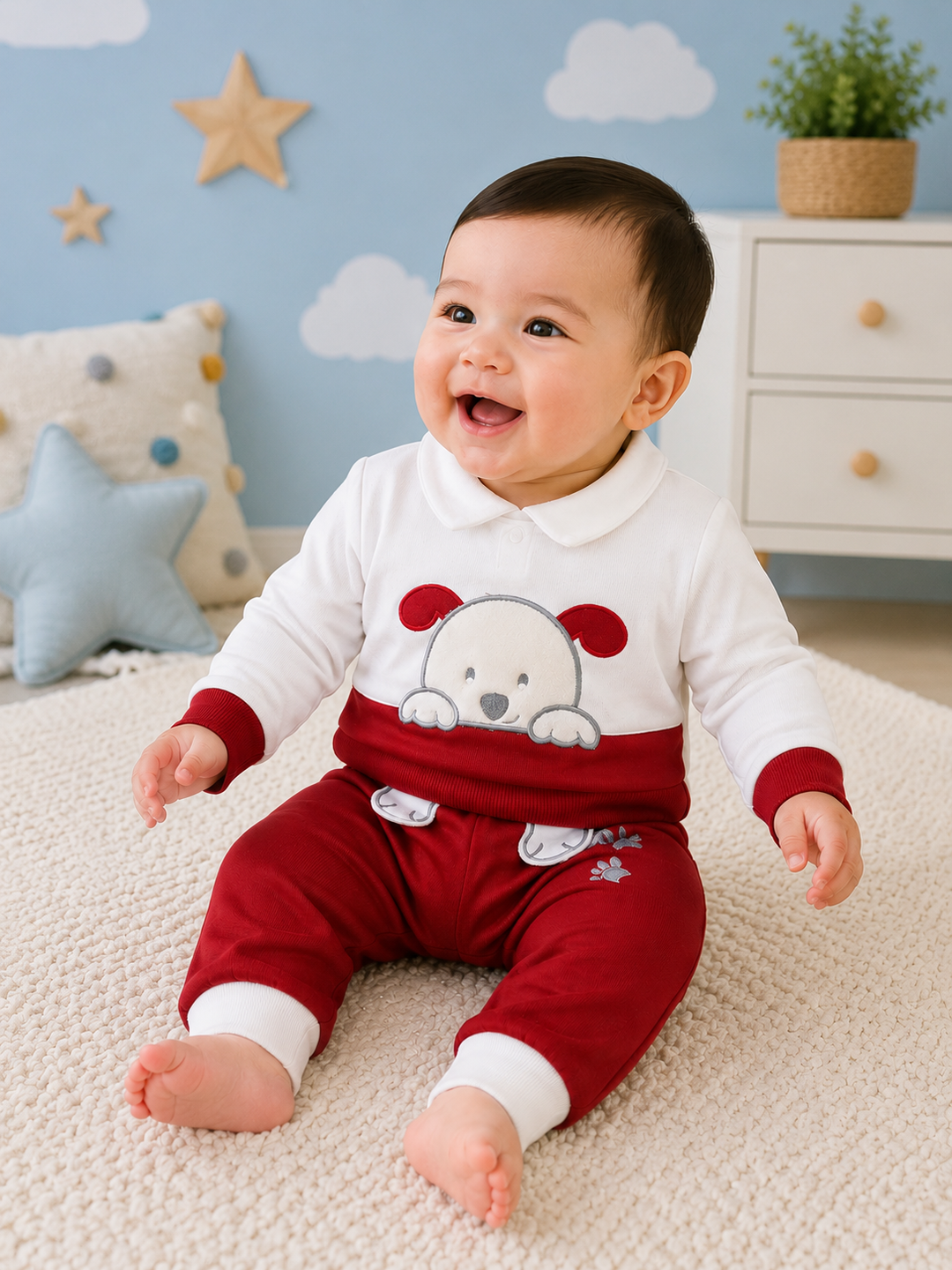 A smiling baby sitting on a cream carpet in a nursery, wearing a white and red long-sleeve romper with a cartoon bear face on the chest.
