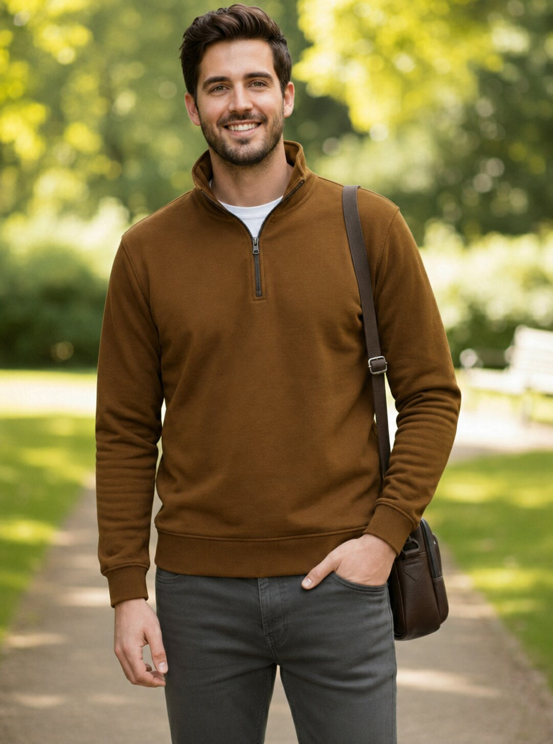 A man wearing a brown quarter-zip pullover sweater over a white t-shirt and gray pants, standing on a park path with a brown shoulder bag.