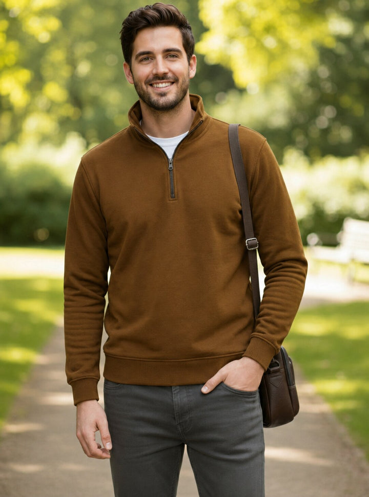 A man wearing a brown quarter-zip pullover sweater over a white t-shirt and gray pants, standing on a park path with a brown shoulder bag.