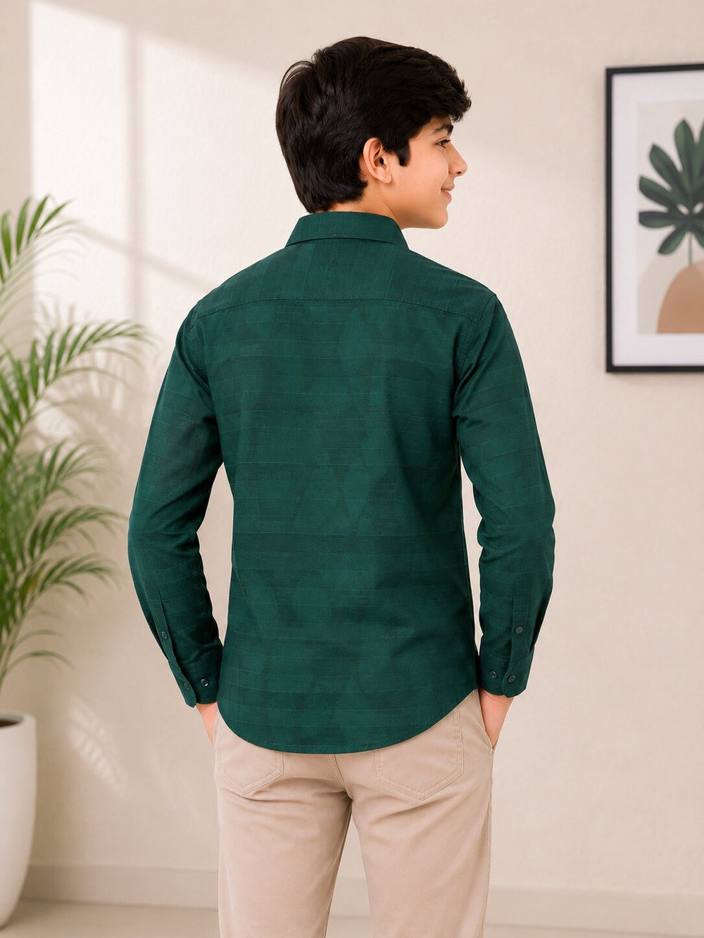 A boy wearing a dark green long-sleeve button-up shirt and beige trousers, shown from the back in a brightly lit indoor setting with a plant and framed wall art.