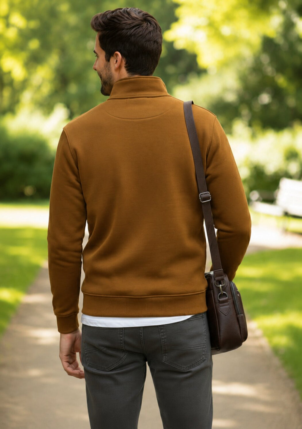 A man viewed from behind wearing a brown mock neck sweater, gray pants, and carrying a brown leather crossbody bag on a sunny path in a park.