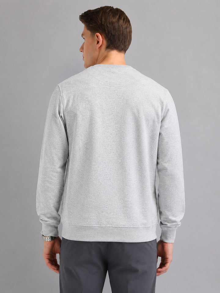 A man viewed from the back wearing a light gray heather crewneck sweatshirt and dark gray trousers, standing against a solid gray studio background.
