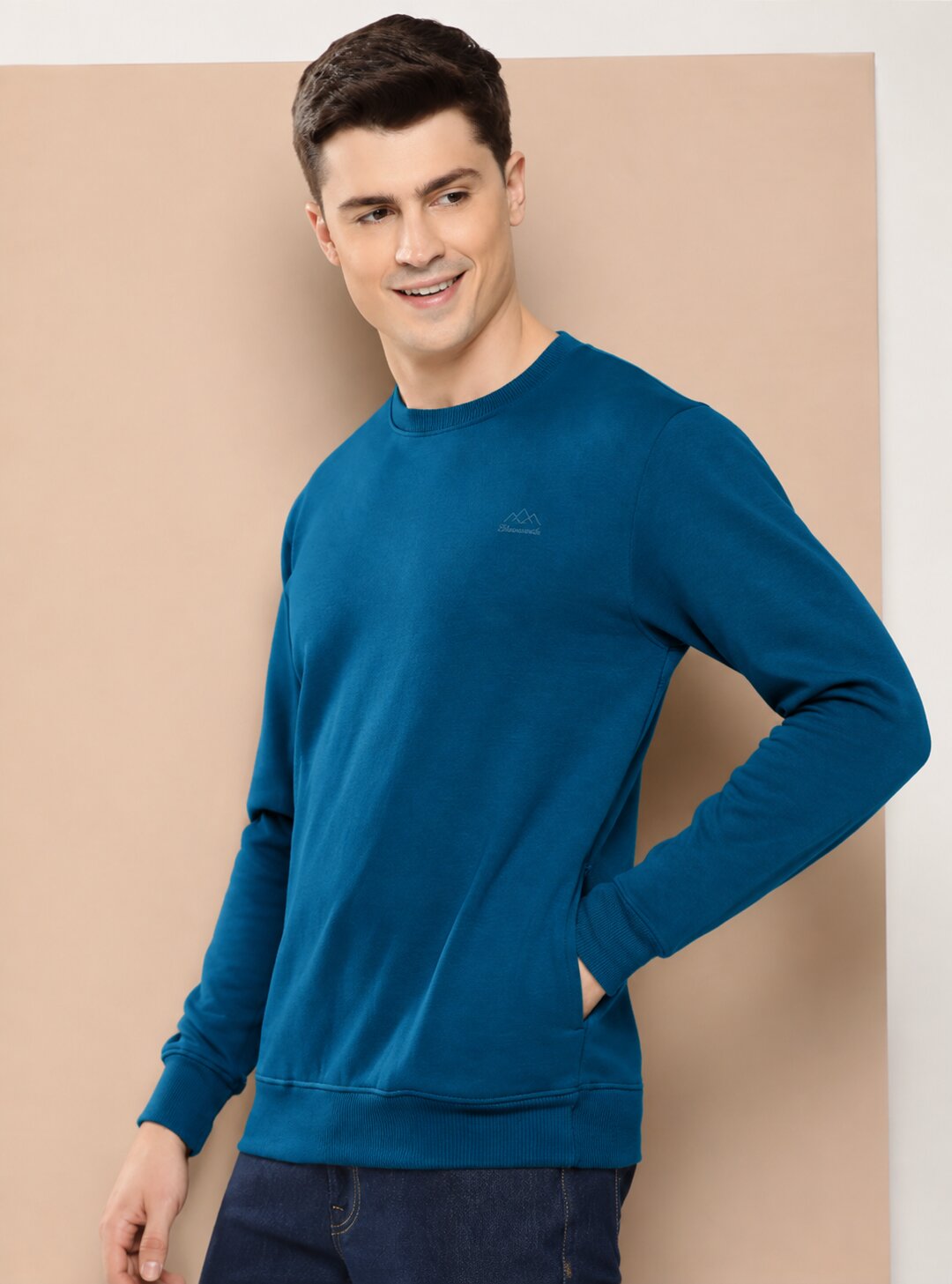 A man wearing a teal blue long-sleeve crewneck sweatshirt with a small embroidered logo on the left chest, paired with dark blue jeans, standing against a beige studio backdrop.