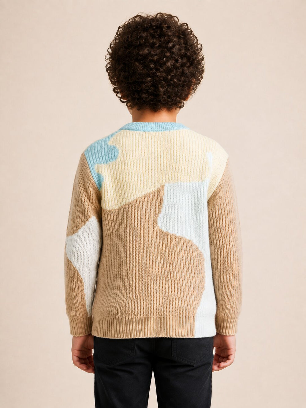 A child viewed from behind wearing a ribbed knit sweater with large abstract color blocks in beige, cream, light blue, and white, paired with dark pants, against a plain beige studio background.