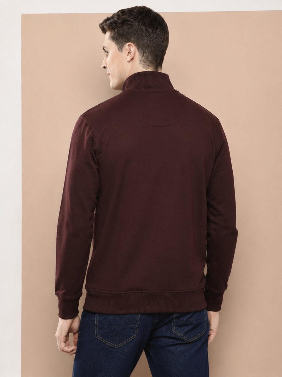 A man viewed from the back wearing a dark maroon long-sleeve mock-neck sweater and blue jeans, standing against a solid beige background.