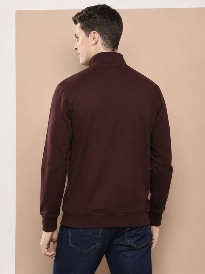 A man viewed from the back wearing a dark maroon long-sleeve mock-neck sweater and blue jeans, standing against a solid beige background.