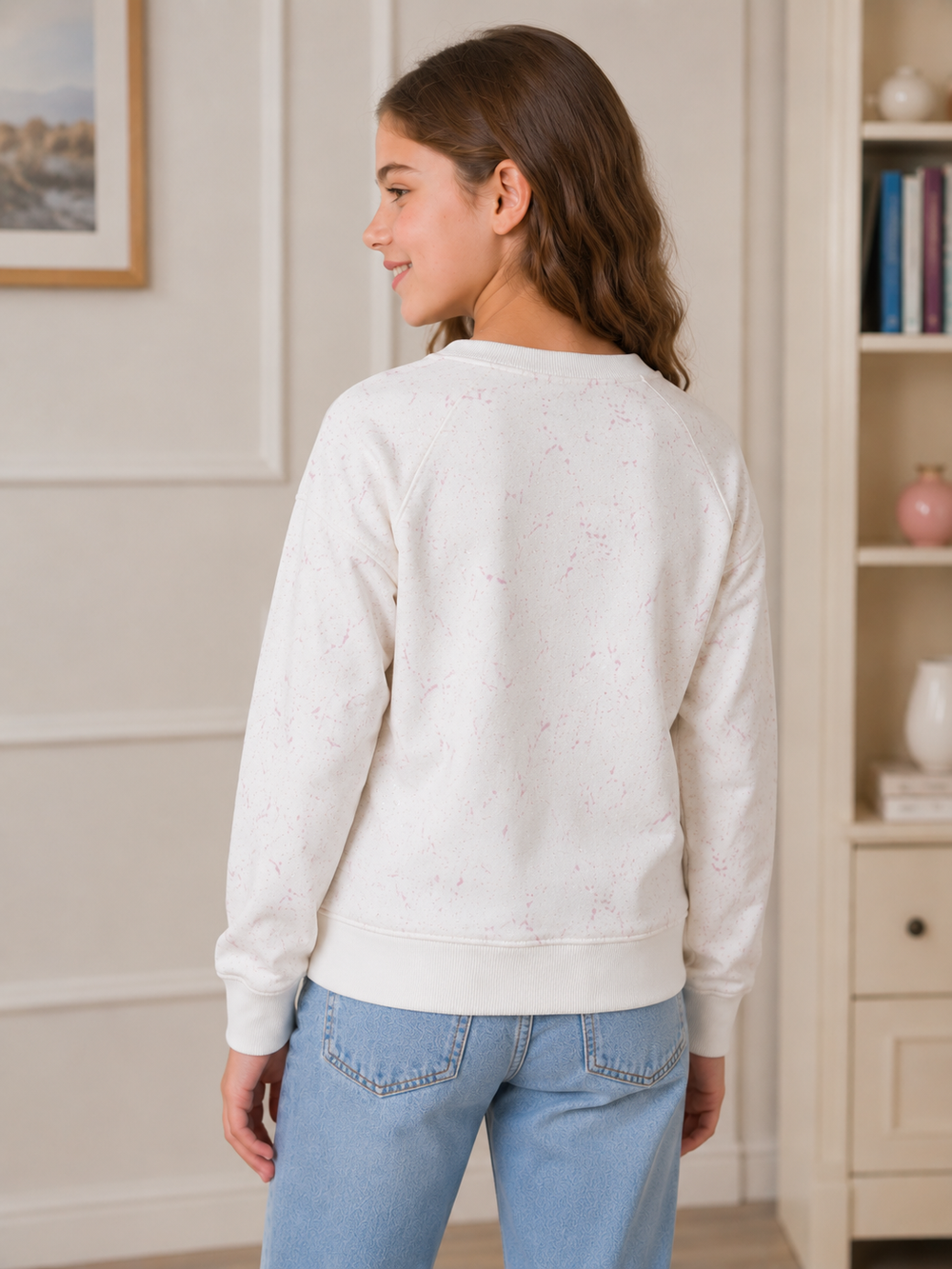 A woman shown from the back wearing a cream-colored crewneck sweatshirt with a subtle pink speckled pattern, paired with light blue jeans, in a bright, neutral-toned room.