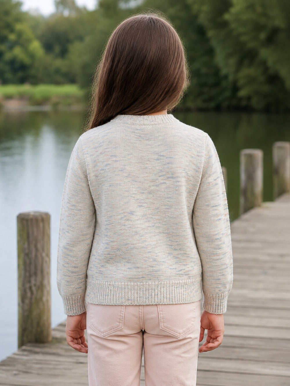 A child viewed from behind wearing a light oatmeal knit sweater and pale pink pants, standing on a wooden dock with water and green trees in the background.
