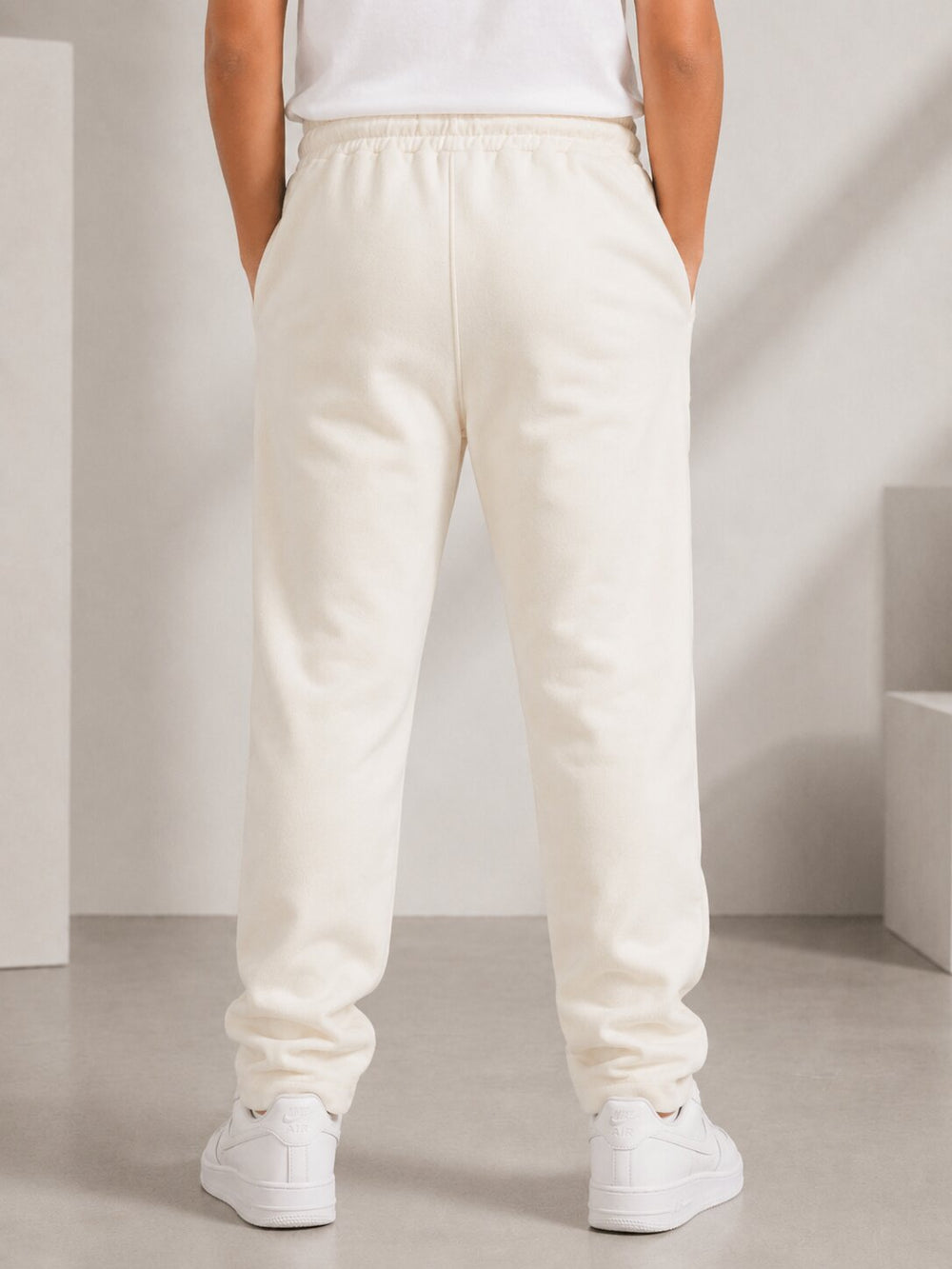 A person shown from the back wearing cream-colored sweatpants, a white top, and white sneakers in a minimalist studio setting.