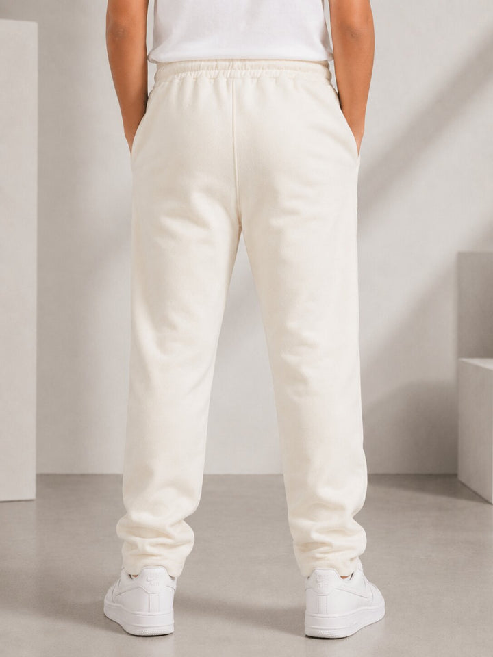 A person shown from the back wearing cream-colored sweatpants, a white top, and white sneakers in a minimalist studio setting.