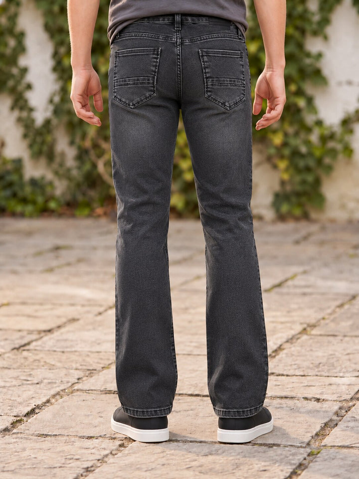 Rear view of a person wearing dark gray washed denim jeans with a straight-to-flare leg, standing on a stone paved surface with green ivy in the background.