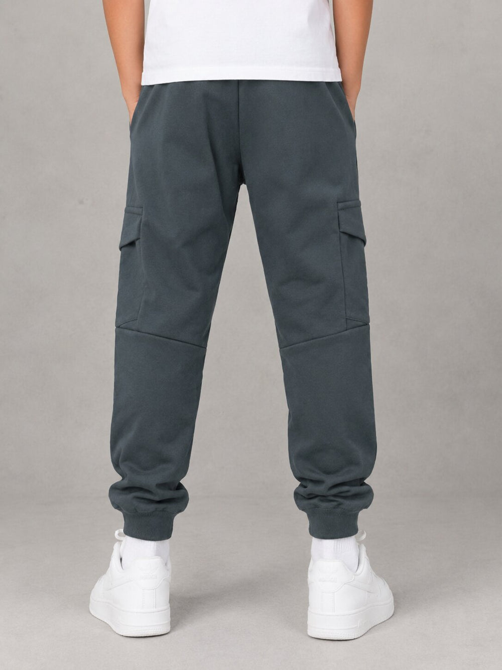 A person shown from the back wearing dark slate gray cargo jogger pants with side flap pockets, a white t-shirt, and white sneakers against a neutral gray studio background.