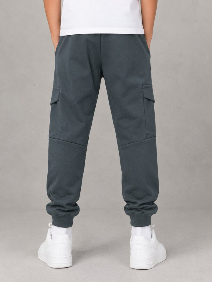 A person shown from the back wearing dark slate gray cargo jogger pants with side flap pockets, a white t-shirt, and white sneakers against a neutral gray studio background.