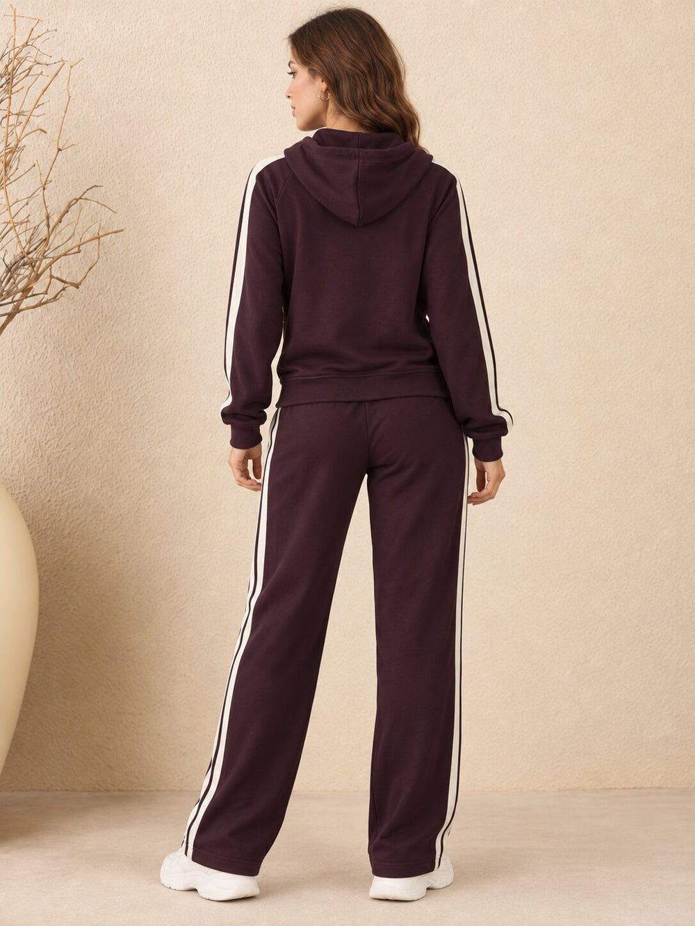 A woman viewed from the back wearing a matching dark plum tracksuit with white side stripes, consisting of a hooded sweatshirt and wide-leg pants, standing against a textured beige wall.