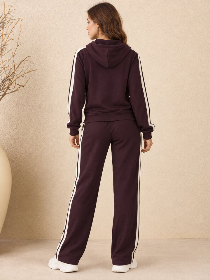 A woman viewed from the back wearing a matching dark plum tracksuit with white side stripes, consisting of a hooded sweatshirt and wide-leg pants, standing against a textured beige wall.