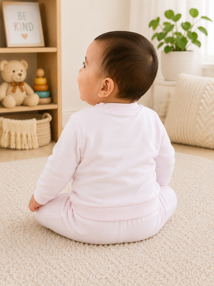 A baby sitting cross-legged on a cream carpet, viewed from behind, wearing a light pink long-sleeve top and matching pink pants in a bright, cozy room with shelves, toys, and a plant.