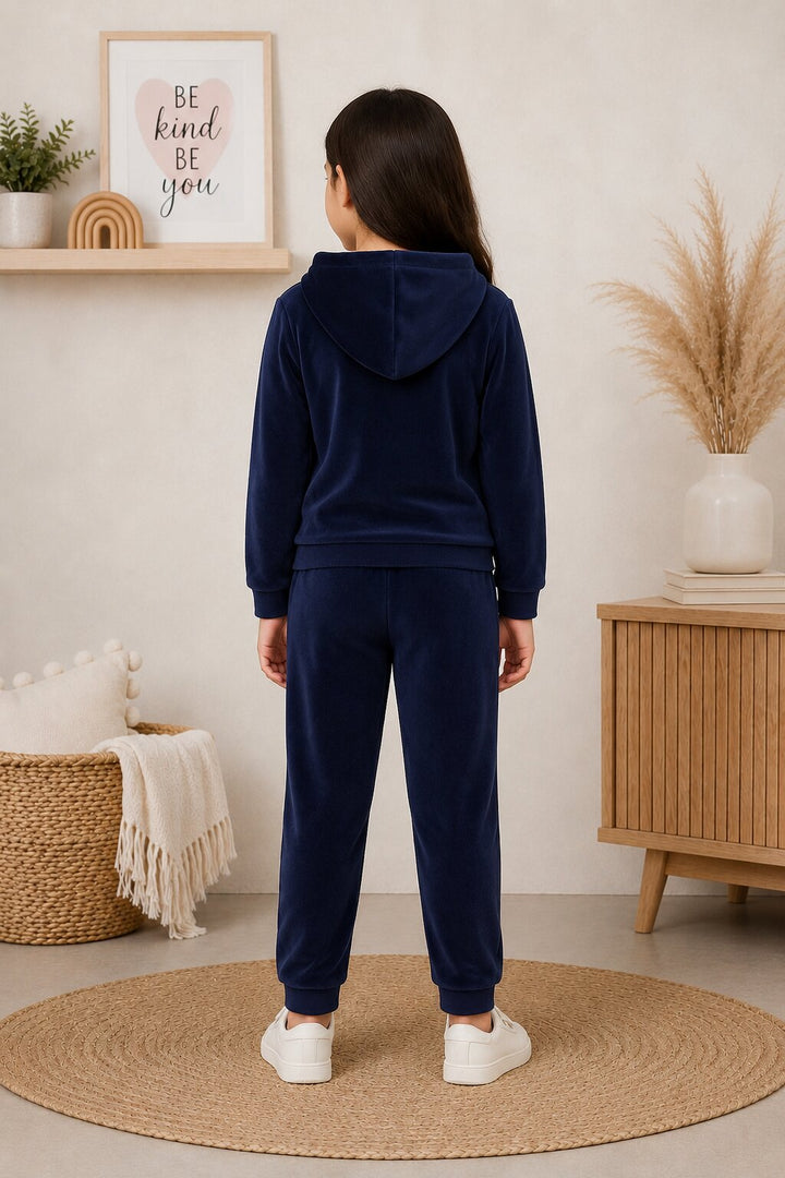 A child stands with their back to the camera wearing a matching navy blue velour loungewear set, consisting of a hooded sweatshirt and jogger pants, in a styled indoor room with a wooden sideboard, pampas grass, and a woven rug.
