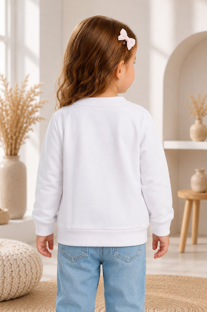 A young girl with brown hair and a white bow is shown from behind wearing a plain white long-sleeve sweatshirt and light blue jeans in a bright, modern indoor room.