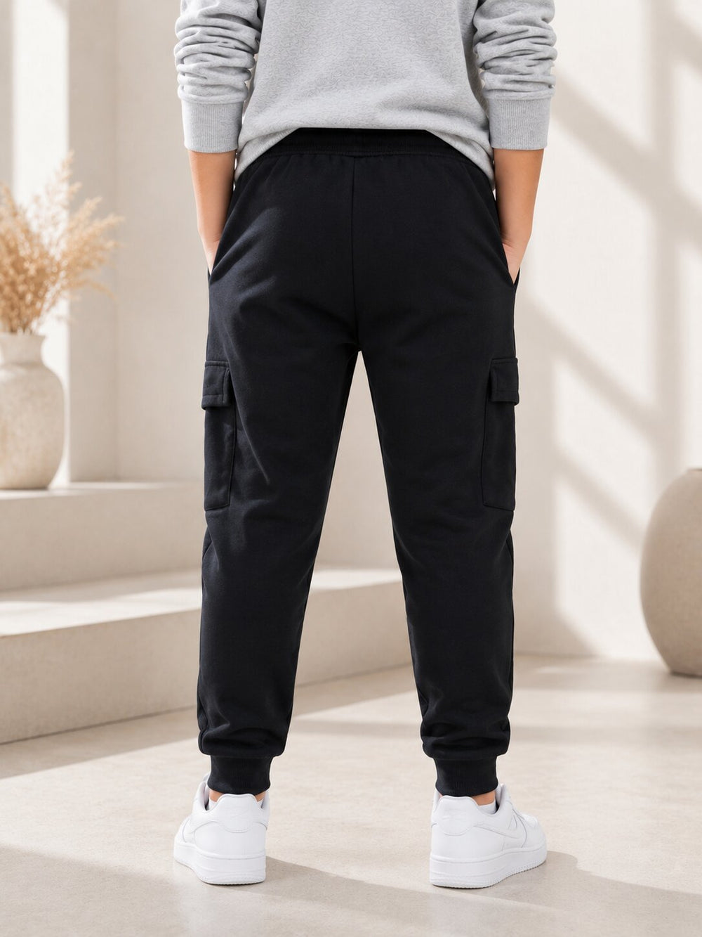 A person shown from the back wearing black cargo jogger pants, a light gray sweatshirt, and white sneakers in a bright, modern indoor setting with beige walls and decorative vases.