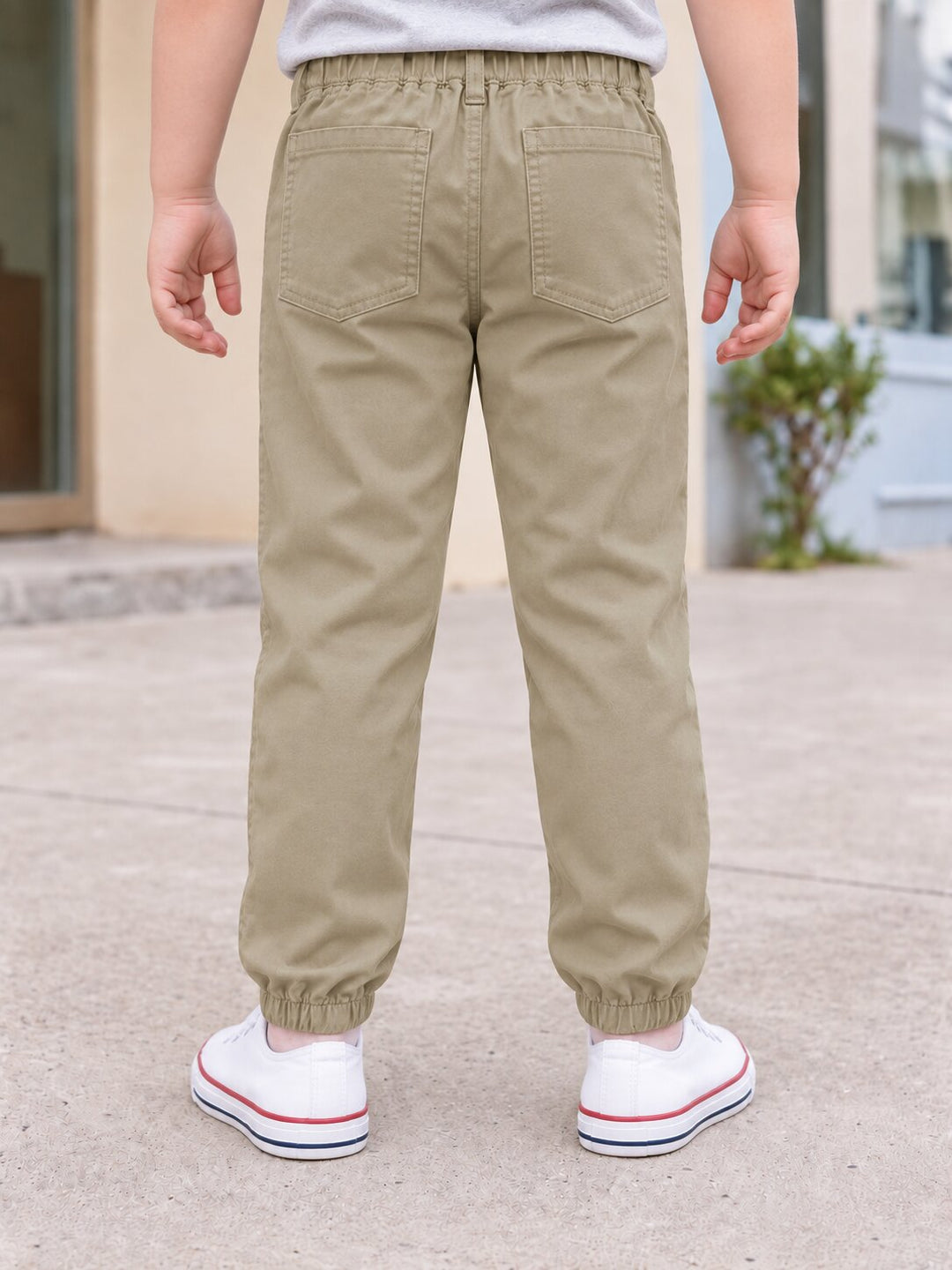 A child shown from the back wearing khaki elastic-waist pants with elastic cuffs and white sneakers, standing outdoors on a paved surface.